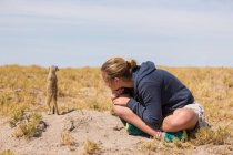 Fille de 12 ans assise à regarder Meerkat émerger du terrier, dans le désert du Kalahari. — Photo de stock