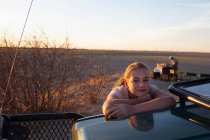 Twelve year old girl leaning on the roof of a vehicle in the Kalahari Desert at sunset. — Stock Photo