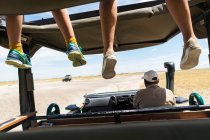 Un veicolo safari, una persona sul sedile di guida e due gambe pendenti dei passeggeri sulla piattaforma di osservazione. — Foto stock