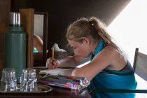 Una niña de doce años sentada en un escritorio en una tienda de campaña en un campamento de reserva de vida silvestre, dibujando. - foto de stock