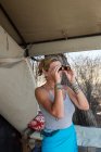 Mature woman by a tent in a wildlife reserve camp using binoculars. — Stock Photo