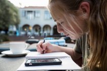 Дівчинка-підліток використовує ручку і папір, робить нотатки, дивлячись на смартфон, пишучи щоденник або роблячи домашнє завдання . — стокове фото