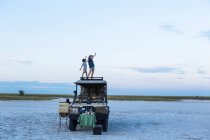 Brother and sister standing on top of a safari vehicle at dusk in a salt pan landscape — Stock Photo