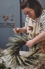 Mujer haciendo una corona de invierno, añadiendo hierbas secas y cabezas de semillero y ramitas con hojas marrones. - foto de stock