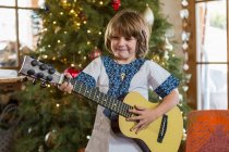 Smiling 4 year old boy playing guitar with Christmas tree in background — Stock Photo