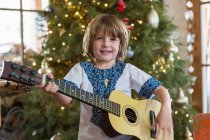 Smiling 4 year old boy playing guitar with Christmas tree in background — Stock Photo