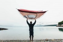 Homme tenant une tente de camping au-dessus de la tête, debout sur une plage rocheuse, une crique sur la côte de l'Alaska. — Photo de stock
