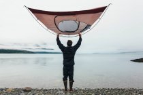Homem segurando barraca de acampamento sobre a cabeça, em pé na praia rochosa, uma entrada na costa do Alasca. — Fotografia de Stock