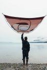 Homem segurando barraca de acampamento sobre a cabeça, em pé na praia rochosa, uma entrada na costa do Alasca. — Fotografia de Stock