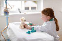 Jeune fille habillée en docteur prétendant traiter le chien jouet dans le lit d'hôpital make-bleieve — Photo de stock