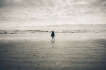 Adolescent garçon debout sur vaste plage, vagues et ciel couvert dans la distance — Photo de stock