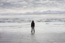 Adolescent garçon debout sur vaste plage, vagues et ciel couvert dans la distance — Photo de stock