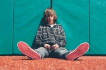 Teenager sitzt auf Sportplatz gegen gepolsterte Wand und hält Handy in der Hand — Stockfoto