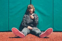 Ragazzo adolescente seduto contro il muro imbottito al campo sportivo, tenendo il telefono cellulare — Foto stock