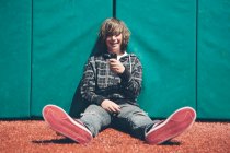 Adolescent garçon assis contre rembourré mur à terrain de sport, tenant téléphone mobile — Photo de stock