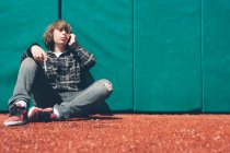 Adolescente sentado contra la pared acolchada en el campo de deportes, sosteniendo el teléfono móvil - foto de stock