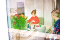 Jeune fille utilisant des peintures à la table de cuisine avec la mère assise à proximité tenant bébé — Photo de stock
