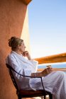 Доросла жінка сидить з напоєм на балконі готелю з видом на блакитний океан і білий піщаний пляж — стокове фото