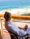 Donna adulta seduta con un drink su un balcone dell'hotel affacciato su un oceano blu e una spiaggia di sabbia bianca — Foto stock