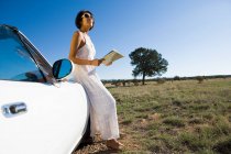 Mulher nativa americana em vestido de sol dirigindo um carro esporte conversível branco na estrada de terra do deserto segurando um mapa — Fotografia de Stock
