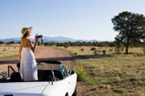 Mujer nativa americana en vestido de sol conduciendo un coche deportivo convertible blanco en el camino de tierra del desierto - foto de stock