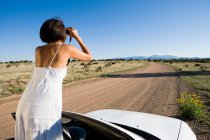 Mulher nativa americana em vestido de sol dirigindo um carro esporte conversível branco na estrada de terra do deserto — Fotografia de Stock