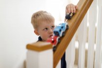 Um menino de três anos brincando com seus carros de brinquedo no corrimão da escada. — Fotografia de Stock