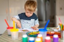 Niño de tres años ocupado pintando en casa, con ollas y pinceles. - foto de stock