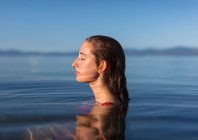 Teenage girl with eyes closed, head and shoulders above the calm waters of a lake at dawn — Stock Photo