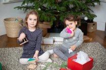 Two young girls sitting on living room floor, unwrapping Christmas present in red box. — Stock Photo