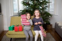 Two young girls sitting on sofa in living room, holding red and white Christmas stocking, smiling at camera. — Stock Photo