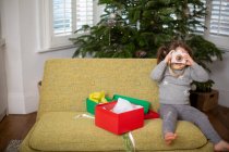 Young girl sitting on sofa in living room with Christmas present in red box, taking picture with toy camera. — Stock Photo