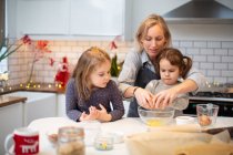 Femme blonde portant tablier bleu et deux filles debout dans la cuisine, la cuisson des biscuits de Noël. — Photo de stock