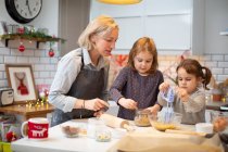 Blond woman wearing blue apron and two girls standing in kitchen, baking Christmas cookies. — Stock Photo