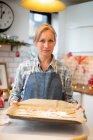 Blonde Frau mit blauer Schürze steht in der Küche, hält Tablett mit Weihnachtsplätzchen in der Hand, blickt in die Kamera. — Stockfoto