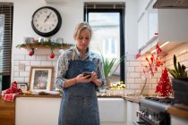 Donna bionda che indossa grembiule blu in piedi in cucina, utilizzando il telefono cellulare. — Foto stock