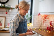 Femme blonde portant tablier bleu debout dans la cuisine, en utilisant un téléphone mobile. — Photo de stock