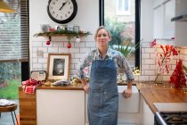 Blonde Frau mit blauer Schürze steht in der Küche und lächelt in die Kamera. — Stockfoto