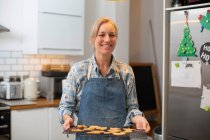 Mujer rubia vistiendo delantal azul de pie en la cocina, sosteniendo bandeja con galletas de Navidad, sonriendo a la cámara. - foto de stock