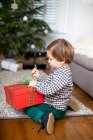 Мальчик сидит на полу в гостиной, разворачивает рождественский подарок в красной коробке. — стоковое фото