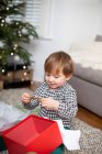 Young boy sitting on living room floor, unwrapping Christmas present in red box. — Stock Photo