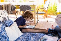 Хлопчик з коричневим волоссям сидить на відкритому ліжку, роблячи домашнє завдання на ноутбуці . — стокове фото