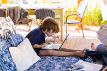 Menino com cabelo castanho sentado na cama ao ar livre, fazendo lição de casa no laptop. — Fotografia de Stock
