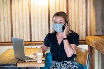 Young blond woman wearing blue face mask, sitting at table, using mobile phone and laptop computer. — Stock Photo