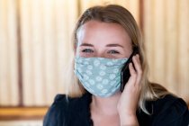 Portrait of young blond woman wearing blue face mask, using mobile phone. — Stock Photo