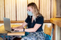 Молода блондинка в синій масці для обличчя, сидить за столом, використовуючи ноутбук . — стокове фото