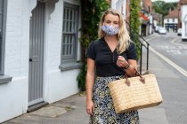 Young blond woman wearing face mask walking through village, carrying shopping bag. — Stock Photo