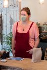 Mujer que usa mascarilla facial y delantal rojo que sirve al cliente en la tienda de productos integrales sin residuos. - foto de stock