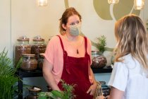 Woman wearing face mask and red apron serving customer in waste free wholefood store. — Stock Photo
