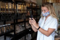 Mulher usando máscara facial em loja local sem resíduos, usando um telefone celular — Fotografia de Stock